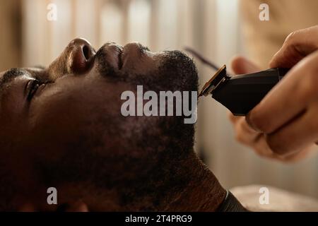 Nahaufnahme eines afroamerikanischen Mannes, der auf einem Stuhl liegt, während der Friseur seinen Bart rasiert Stockfoto