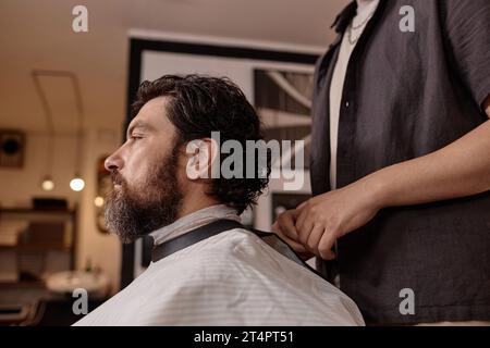Reifer Mann sitzt im Stuhl, während er sich im Friseur Haare und Bart schneidet Stockfoto