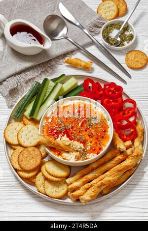 Heißer Pfeffer Gelee Käse Dip in Schüssel serviert mit Crackern, salzigen grissini, frischem Pfeffer und Gurke auf Platte auf weißem Holztisch, vertikale Ansicht Stockfoto