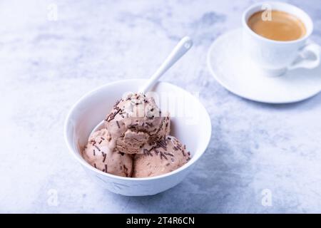 Schokoladeneis mit Schokolade in einer weißen Schüssel auf einem hellen Hintergrund streuen. Drei Kugeln hausgemachtes Eis und eine Tasse Kaffee. Nahaufnahme. Stockfoto