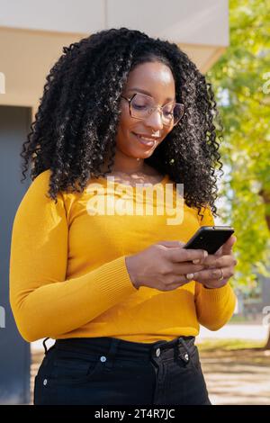 Vertikale Aufnahme Junge glückliche Afroamerikanerin, die im Internet mit intelligenter mobiler Anwendung im Freien surft. Stockfoto