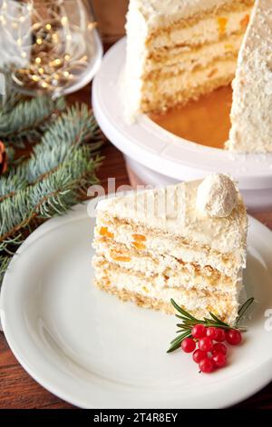 Ein Stück festlicher Weihnachtskuchen mit Kokos und Pfirsichen, dekoriert mit Preiselbeeren und Rosmarin auf weißem Teller. Stockfoto