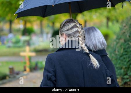 Nahaufnahme der Tochter, die ihre Mutter in den Armen hält, auf dem Weg zum Grab des Mannes auf dem Friedhof an einem wolkigen kalten Tag Stockfoto