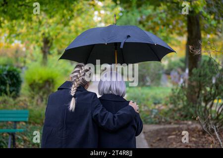 Nahaufnahme der Tochter, die ihre Mutter in den Armen hält, auf dem Weg zum Grab des Mannes auf dem Friedhof an einem wolkigen kalten Tag Stockfoto