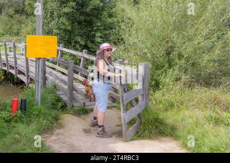 Reifer Wanderer zusammen mit seinem Hund öffnet und überquert das Tor zur Brücke, die zum Naturschutzgebiet führt, wilde Vegetation im Hintergrund, Hut und Sonnenbrille Stockfoto
