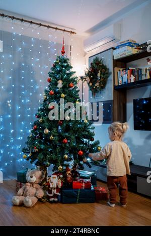 Das kleine Mädchen steht neben einem geschmückten Weihnachtsbaum und berührt die Bälle. Rückansicht Stockfoto