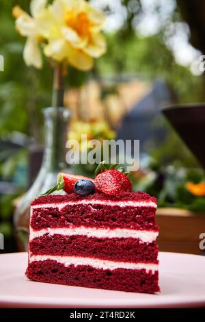 Ein Stück roter Samtkuchen, dekoriert mit frischer Erdbeere und Heidelbeere auf rosa Teller im Café im Freien. Stockfoto