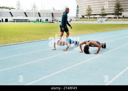 Halten Halten Sie es. Langaufnahme von zwei attraktiven jungen Athleten, die auf einem Rennfeld Pressen machen, während ihr Trainer zusieht. Stockfoto