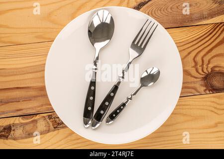 Zwei Metalllöffel und Gabel mit weißer Keramikplatte auf Holztisch, Makro, Draufsicht. Stockfoto