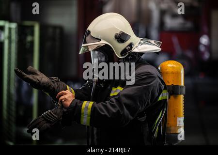 Feuerwehrporträt mit voller Ausrüstung und Sauerstoffmaske. Das Anziehen von Schutzhandschuhen. Feuerwehrfahrzeuge im Hintergrund. Stockfoto