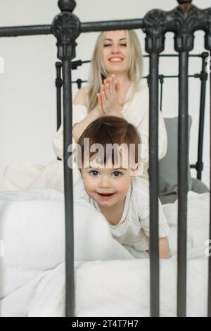 Porträt eines kleinen Mädchens, das, aufgewacht, mit ihrer Mutter im Bett spielt, vor ihr kriecht und lächelt. Familienkonzept. Vertikaler Rahmen. Stockfoto