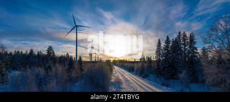 Panoramaaufnahme über die Winterstraße im Wald mit Windmühlen, die auf der linken Seite im Wald stehen. Straße auf der rechten Seite Stockfoto