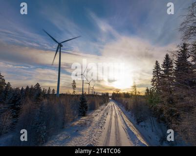 Landschaftlich reizvolle Luftaufnahme über der Waldwinterstraße mit Windmühlen, die auf der linken Seite im Wald stehen. Straße in der Mitte der Sicht. Große Windturbinen Stockfoto