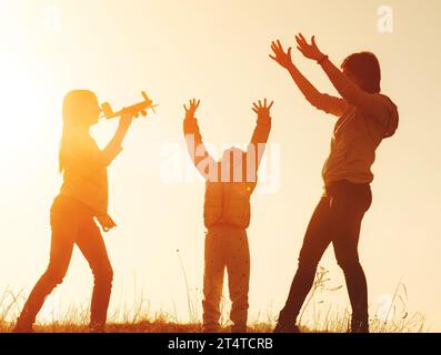 Mutter mit zwei kleinen Töchtern, die Spaß bei Sonnenuntergang haben. Silhouette von Mutter und ihren jungen Töchtern, die mit Flugzeugspielzeug auf dem Grasfeld spielen. Glückliches Familienkonzept Stockfoto