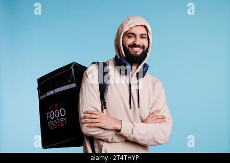 Arabischer Mann mit fröhlichem Ausdruck, der Rucksack trägt und ein Porträt liefert. Junger, lächelnder Restaurantlieferant mit Kapuze, stehend mit Thermotasche und Blick in die Kamera Stockfoto