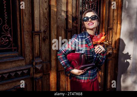 Porträt einer stilvollen Frau, die burgunderrote Geldbörse hält, die karierte Blazer rote Kleid Sonnenbrille auf der Stadtstraße durch Holztüren trägt. Herbstmodisch alt Stockfoto