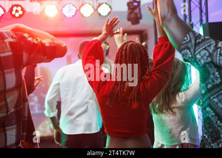 Leute tanzen und heben Hände, während sie an einer dj-Party im Nachtclub teilnehmen. Clubfans mit Fans und Springen auf der überfüllten Tanzfläche in der Diskothek im Club, die von lebhaften Lichtern beleuchtet ist Stockfoto