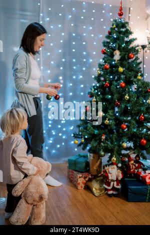 Ein kleines Mädchen mit einem Teddybären in der Hand sieht zu, wie ihre Mutter den Weihnachtsbaum schmückt. Rückansicht Stockfoto