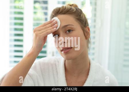 Glückliche kaukasierin im Bademantel, der Gesicht mit Wattepad im sonnigen Badezimmer reinigt Stockfoto
