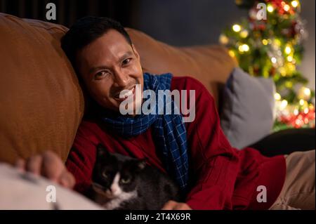 Ein glücklicher asiatischer Mann liegt mit seiner lieblichen Katze auf einem Sofa und feiert gemeinsam Weihnachten zu Hause. Hauskatze und Besitzer zu Hause. Haustierkonzept Stockfoto