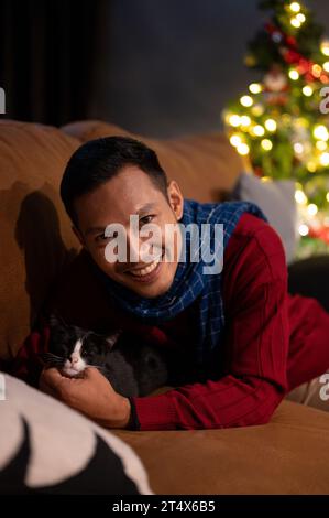 Ein glücklicher asiatischer Mann liegt mit seiner lieblichen Katze auf einem Sofa und feiert gemeinsam Weihnachten zu Hause. Hauskatze und Besitzer zu Hause. Haustierkonzept Stockfoto