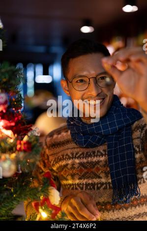 Ein Porträt eines glücklichen, gutaussehenden asiatischen Mannes in einem bequemen Schal und Pullover bewundert ein wunderschönes Weihnachtsschmuck, der seinen Weihnachtsbaum im Th schmückt Stockfoto