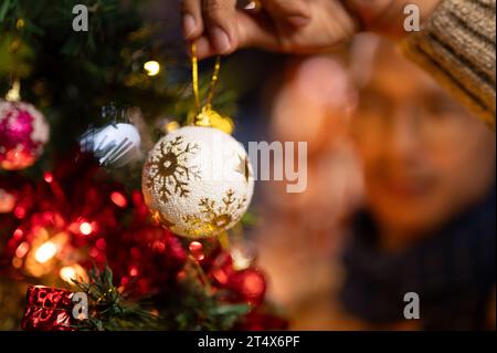 Nahaufnahme eines Mannes, der ein schönes Weihnachtsschmuck hält und im Wohnzimmer ein Weihnachtskugelschmuck an einen Weihnachtsbaum hängt. Stockfoto