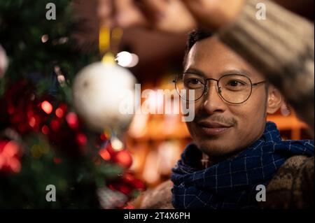 Ein Porträt eines glücklichen, gutaussehenden asiatischen Mannes in einem bequemen Schal und Pullover bewundert ein wunderschönes Weihnachtsschmuck, hängende Ornamente und dekoratives Hi Stockfoto