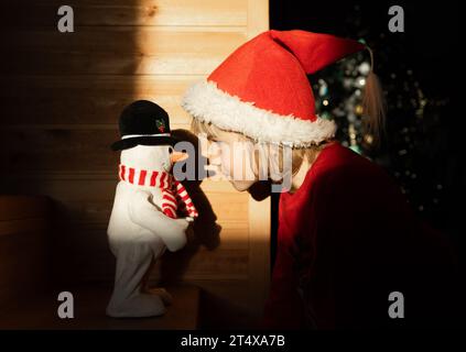 Fröhlicher Junge mit Weihnachtsmann-Hut macht Spaß, mit einem Schneemann von einem Spielzeug Nase an Nase zu drücken. Heiligabend, spielen zu Hause in Erwartung der holi Stockfoto