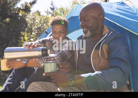 Glücklicher afroamerikanischer Vater und erwachsener Sohn sitzen vor dem Zelt und trinken Kaffee in der Sonne Stockfoto