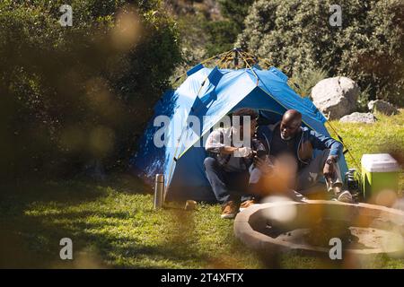 Glücklicher afroamerikanischer Vater und erwachsener Sohn sitzen außerhalb des Zeltes mit Smartphone, Kopierraum Stockfoto