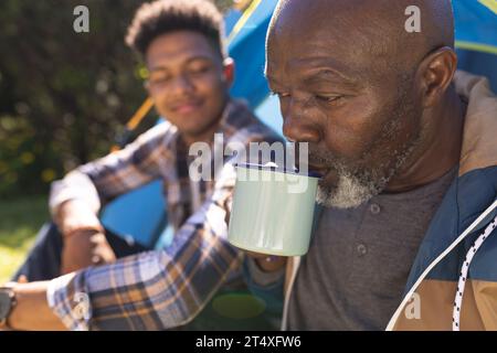 Glücklicher afroamerikanischer Vater, der vor dem Zelt sitzt und Kaffee mit einem erwachsenen Sohn in der Sonne trinkt Stockfoto