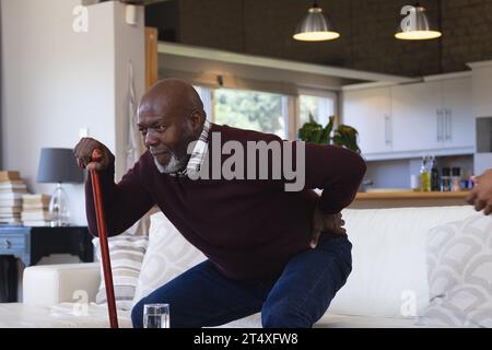 Schmerzhafter afroamerikanischer Senior Mann mit Gehstock, der von der Couch zu Hause aufsteht und sich zurückhält Stockfoto