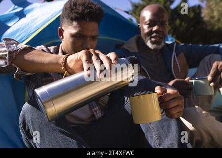 afroamerikanischer Vater und erwachsener Sohn sitzen vor dem Zelt und trinken Kaffee in der Sonne Stockfoto