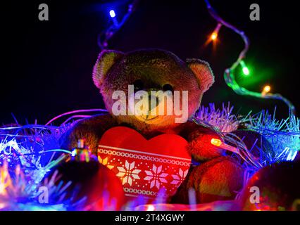 Ein Teddybär sitzt mit einem Weihnachtsbaumspielzeug in Form eines Herzens auf einem festlichen Lametta, umgeben von Weihnachtsdekorationen und bunten Girlanden Stockfoto