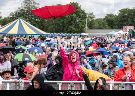 Fans singen bei einem großen Musikfestival in einem Park in Essex bei starkem Regen mit. Eine Frau, die die Show genießt, obwohl ihr Regenschirm sich von innen nach außen dreht Stockfoto