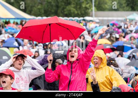 Fans singen bei einem großen Musikfestival in einem Park in Essex bei starkem Regen mit. Frau, die die Show unter ihrem Schirm genießt. Andere in wasserdichten Materialien Stockfoto