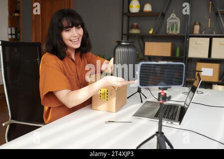 Junge lächelnde brünette Frau, die beim Livestream vor der Smartphone-Kamera handgefertigte Vase vor dem Online-Publikum zeigt Stockfoto