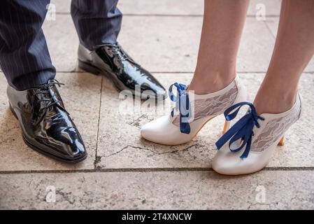 Elegante Herrenschuhe aus schwarzem Lackleder und weiße Damenschuhe mit blauen Schnürsenkeln und Spitze, getragen und gegenüberliegend Stockfoto