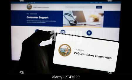 Person mit Handy und Logo der California Public Utilities Commission (CPUC) vor der Webseite. Schwerpunkt auf dem Display des Telefons. Stockfoto