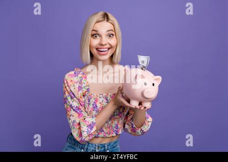 Porträt einer netten, fröhlichen Dame, die Lächeln ausstrahlt, Arme halten Geld Bank Schwein Dollar Scheine Einsparungen isoliert auf lila Hintergrund Stockfoto