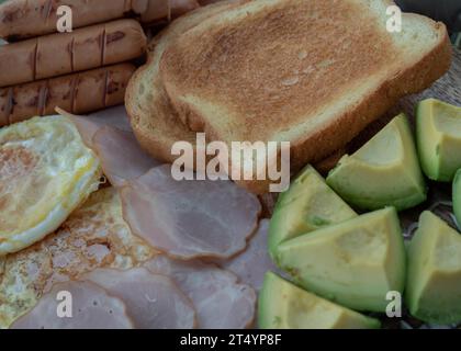 Nahaufnahme des Frühstücks umfasst gebratene Würstchen, gebratene Eier, Toastbrot und geschnittene Avocados. Platz für Text, selektiver Fokus. Stockfoto