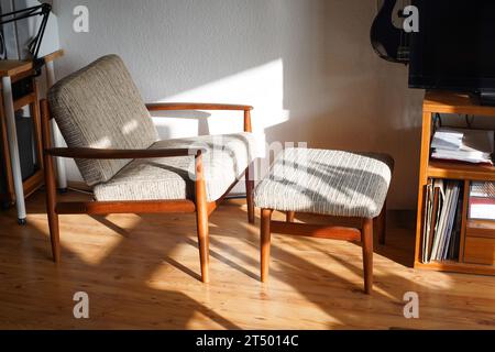 Ein Mid Century Sessel mit Ottoman aus Teakholz im dänischen Design in einem gemütlichen, modern eingerichteten Wohnzimmer im Stil der 60er Jahre mit weißer Wand und Fernseher Stockfoto