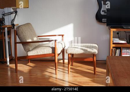 Ein Mid Century Sessel mit Ottoman aus Teakholz im dänischen Design in einem gemütlichen, modern eingerichteten Wohnzimmer im Stil der 60er Jahre mit weißer Wand und Fernseher Stockfoto