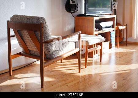 Ein Mid Century Sessel mit Ottoman aus Teakholz im dänischen Design in einem gemütlichen, modern eingerichteten Wohnzimmer im Stil der 60er Jahre mit weißer Wand und Fernseher Stockfoto