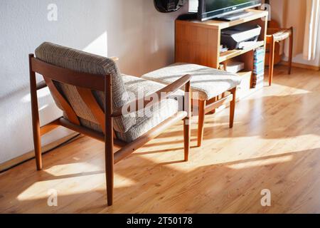 Ein Mid Century Sessel mit Ottoman aus Teakholz im dänischen Design in einem gemütlichen, modern eingerichteten Wohnzimmer im Stil der 60er Jahre mit weißer Wand und Fernseher Stockfoto