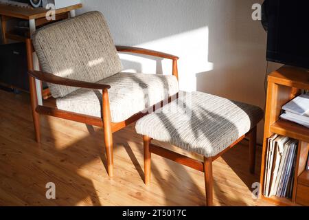 Ein Mid Century Sessel mit Ottoman aus Teakholz im dänischen Design in einem gemütlichen, modern eingerichteten Wohnzimmer im Stil der 60er Jahre mit weißer Wand und Fernseher Stockfoto