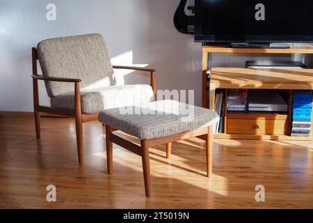 Ein Mid Century Sessel mit Ottoman aus Teakholz im dänischen Design in einem gemütlichen, modern eingerichteten Wohnzimmer im Stil der 60er Jahre mit weißer Wand und Fernseher Stockfoto