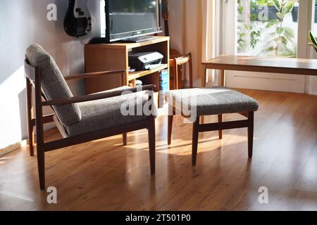 Ein Mid Century Sessel mit Ottoman aus Teakholz im dänischen Design in einem gemütlichen, modern eingerichteten Wohnzimmer im Stil der 60er Jahre mit weißer Wand und Fernseher Stockfoto