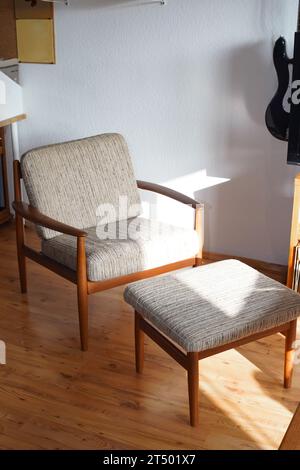 Ein Mid Century Sessel mit Ottoman aus Teakholz im dänischen Design in einem gemütlichen, modern eingerichteten Wohnzimmer im Stil der 60er Jahre mit weißer Wand und Fernseher Stockfoto